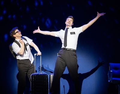 Two performers in white shirts, black ties, and name tags engage in a theatrical scene, one with luggage, on a dark stage with blue lighting.