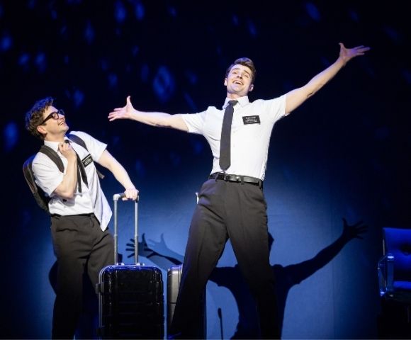 Two performers in white shirts, black ties, and name tags engage in a theatrical scene, one with luggage, on a dark stage with blue lighting.