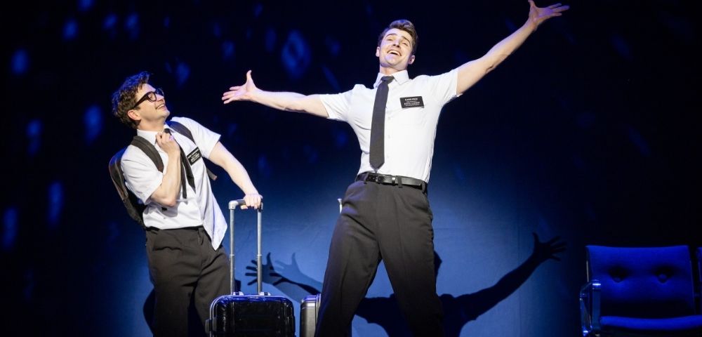 Two performers in white shirts, black ties, and name tags engage in a theatrical scene, one with luggage, on a dark stage with blue lighting.
