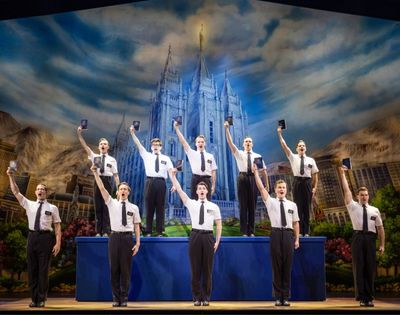 A group of nine men in white shirts and black ties stand on stage holding up black books, with a large illuminated castle backdrop behind them.