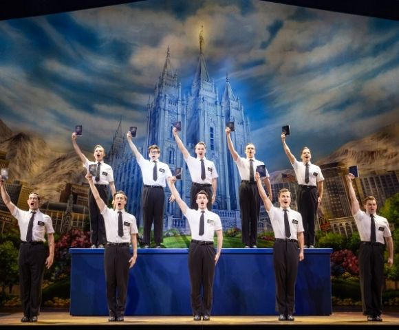 A group of nine men in white shirts and black ties stand on stage holding up black books, with a large illuminated castle backdrop behind them.