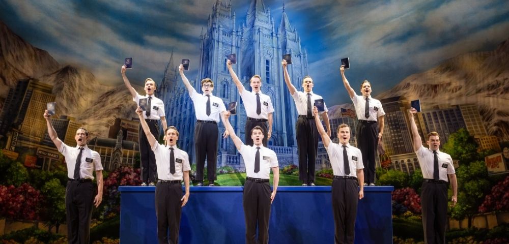 A group of nine men in white shirts and black ties stand on stage holding up black books, with a large illuminated castle backdrop behind them.