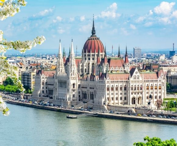 The image shows the Budapest Parliament Building, with Gothic architecture, along the Danube River under a bright blue sky. Blossoming trees frame the scene.