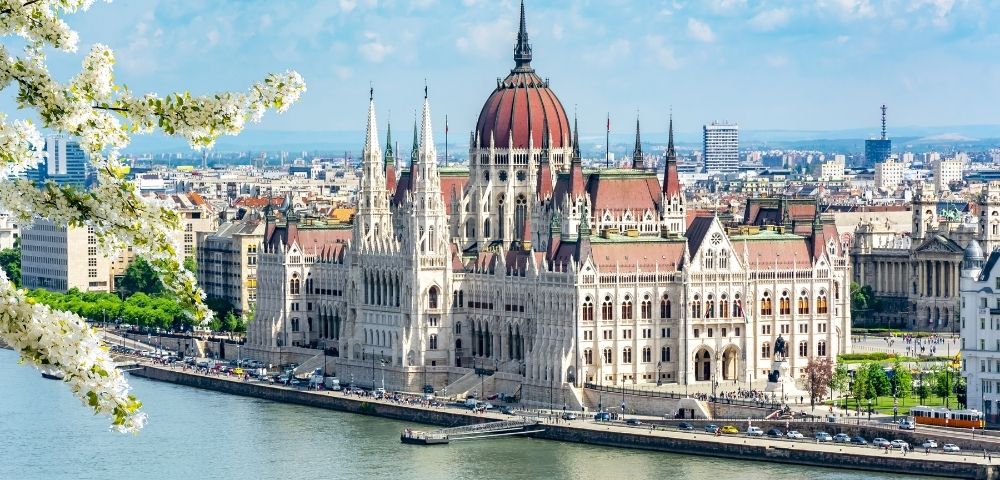 The image shows the Budapest Parliament Building, with Gothic architecture, along the Danube River under a bright blue sky. Blossoming trees frame the scene.