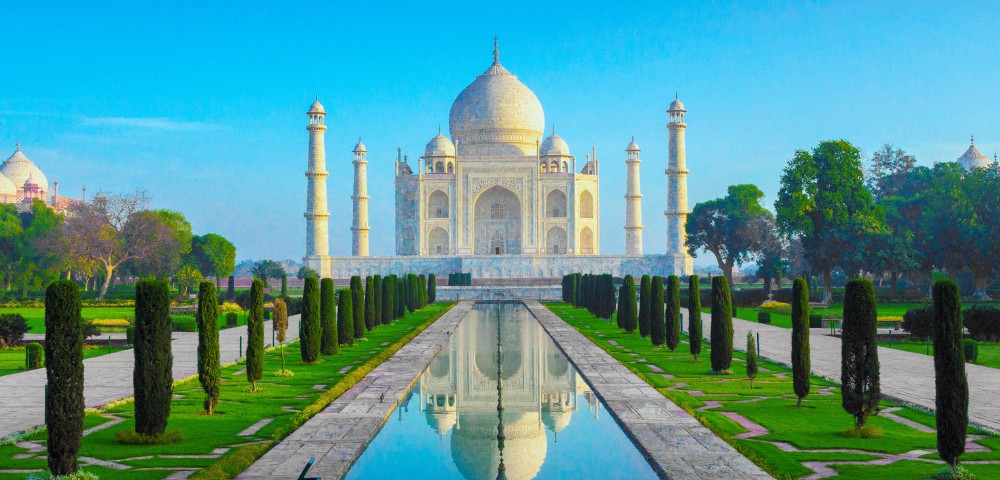 The Taj Mahal, a white marble mausoleum, with its reflection in a long, serene pool. Flanking pathways are lined with lush green trees.