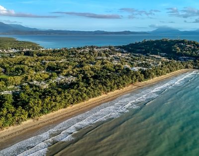 Aerial view of a tropical coastline with a sandy beach, lush green vegetation, and calm turquoise water, leading to a forested headland with scattered buildings.