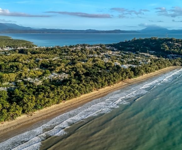Aerial view of a tropical coastline with a sandy beach, lush green vegetation, and calm turquoise water, leading to a forested headland with scattered buildings.