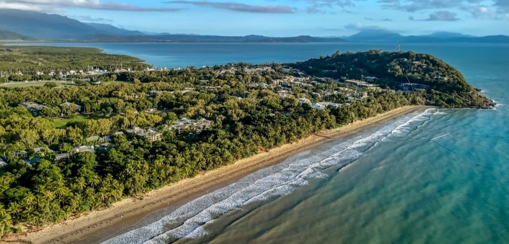Aerial view of a tropical coastline with a sandy beach, lush green vegetation, and calm turquoise water, leading to a forested headland with scattered buildings.
