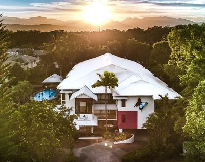 Aerial view of a modern resort building with a white roof surrounded by lush green trees during a sunset with mountains in the background.