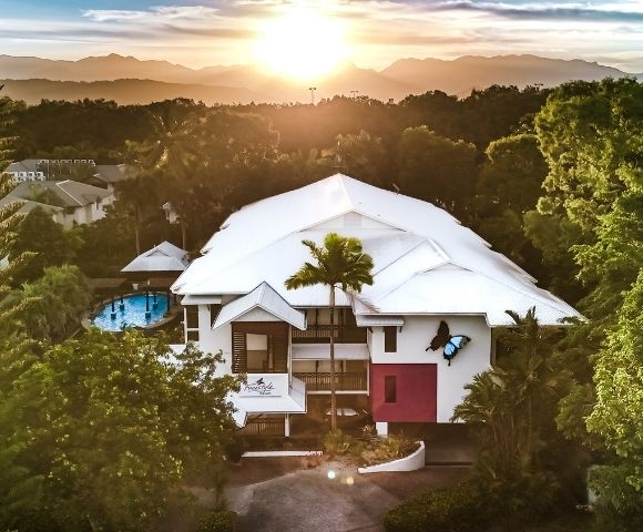 Aerial view of a modern resort building with a white roof surrounded by lush green trees during a sunset with mountains in the background.