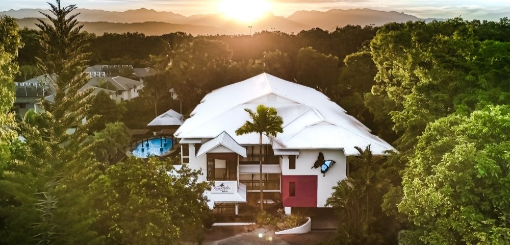 Aerial view of a modern resort building with a white roof surrounded by lush green trees during a sunset with mountains in the background.