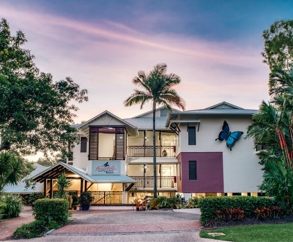 A resort building with a large blue butterfly decoration on the exterior, surrounded by lush greenery and palm trees, under a soft twilight sky.