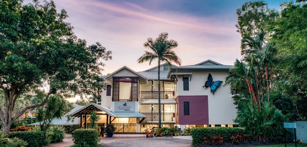 A resort building with a large blue butterfly decoration on the exterior, surrounded by lush greenery and palm trees, under a soft twilight sky.