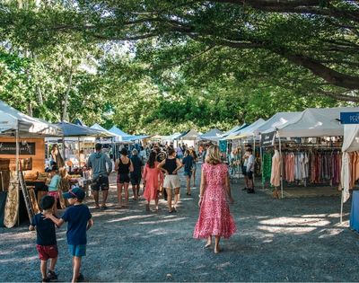 A bustling outdoor market with rows of white tents displaying clothing and crafts, set under a canopy of lush green trees.