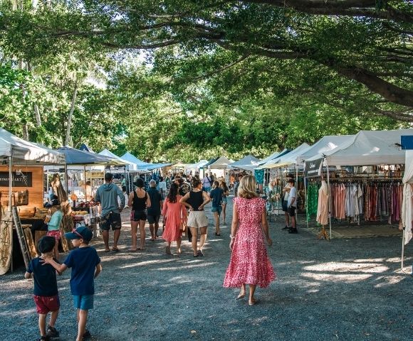 A bustling outdoor market with rows of white tents displaying clothing and crafts, set under a canopy of lush green trees.