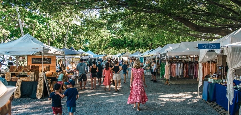 A bustling outdoor market with rows of white tents displaying clothing and crafts, set under a canopy of lush green trees.