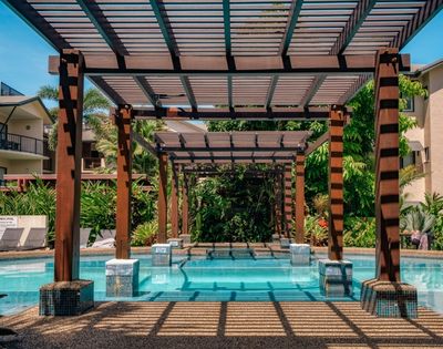 A swimming pool with clear blue water passes under a wooden pergola with a slatted roof, surrounded by lush green foliage and modern apartment buildings on a sunny day.