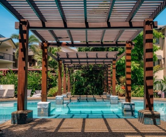 A swimming pool with clear blue water passes under a wooden pergola with a slatted roof, surrounded by lush green foliage and modern apartment buildings on a sunny day.