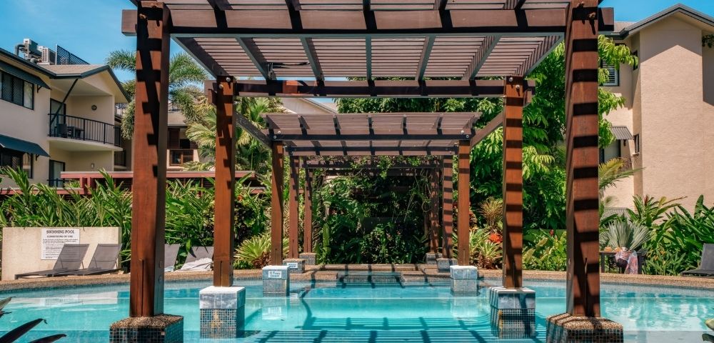 A swimming pool with clear blue water passes under a wooden pergola with a slatted roof, surrounded by lush green foliage and modern apartment buildings on a sunny day.