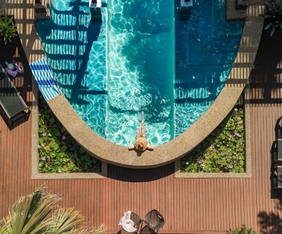 Aerial view of a person relaxing in a swimming pool with steps and water lilies, surrounded by a wooden deck and lounge chairs.