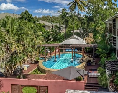 Resort swimming pool area surrounded by lush tropical vegetation and modern apartment buildings, featuring a covered seating area over the water.