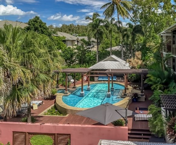 Resort swimming pool area surrounded by lush tropical vegetation and modern apartment buildings, featuring a covered seating area over the water.