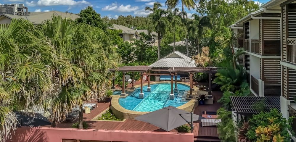 Resort swimming pool area surrounded by lush tropical vegetation and modern apartment buildings, featuring a covered seating area over the water.