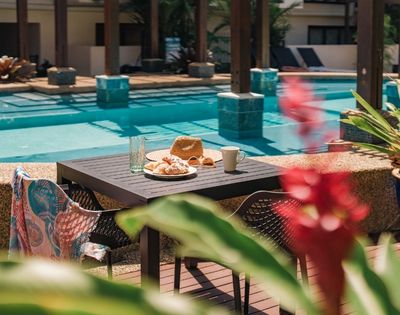 Table set with croissants, a straw hat, sunglasses, a coffee mug, and a glass of water next to a swimming pool.