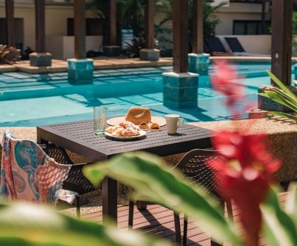 Table set with croissants, a straw hat, sunglasses, a coffee mug, and a glass of water next to a swimming pool.