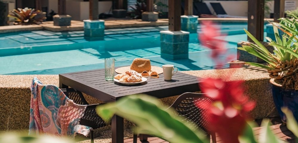 Table set with croissants, a straw hat, sunglasses, a coffee mug, and a glass of water next to a swimming pool.