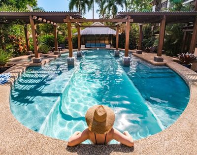 A person in a straw hat sits with their back to the camera, at the edge of a curving, shallow swimming pool with submerged steps. The pool is surrounded by a light-colored pebble-crete deck and lush tropical foliage. A wooden pergola with a slatted roof casts dappled shadows across the bright turquoise water. In the background, a small, open-sided pavilion with blue seating is visible.