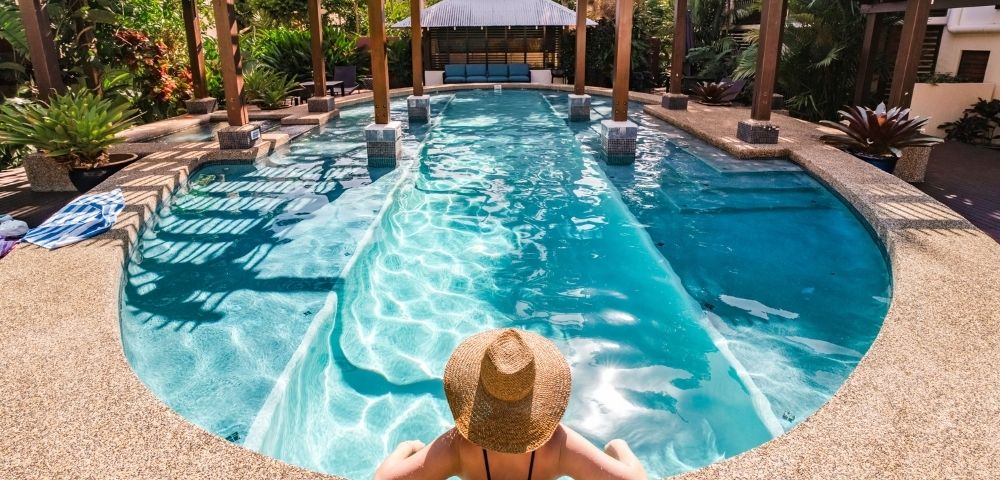 A person in a straw hat sits with their back to the camera, at the edge of a curving, shallow swimming pool with submerged steps. The pool is surrounded by a light-colored pebble-crete deck and lush tropical foliage. A wooden pergola with a slatted roof casts dappled shadows across the bright turquoise water. In the background, a small, open-sided pavilion with blue seating is visible.