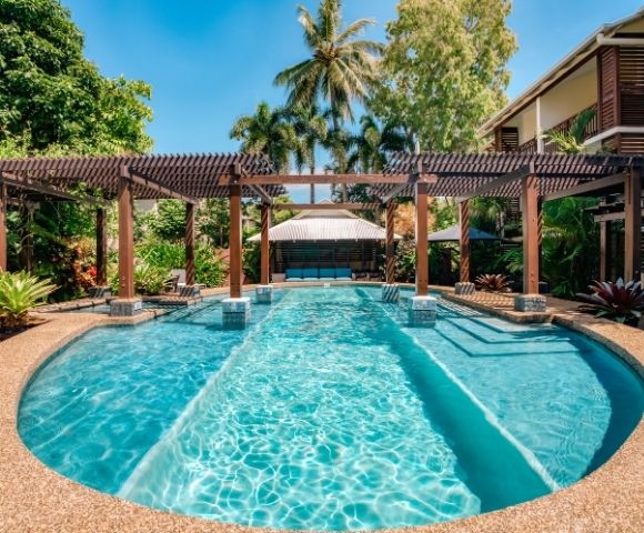 Resort swimming pool with clear turquoise water, surrounded by tropical foliage and a wooden pergola, with a covered seating area in the background.