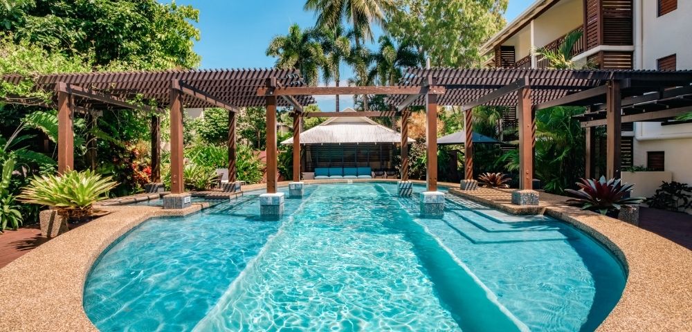 Resort swimming pool with clear turquoise water, surrounded by tropical foliage and a wooden pergola, with a covered seating area in the background.