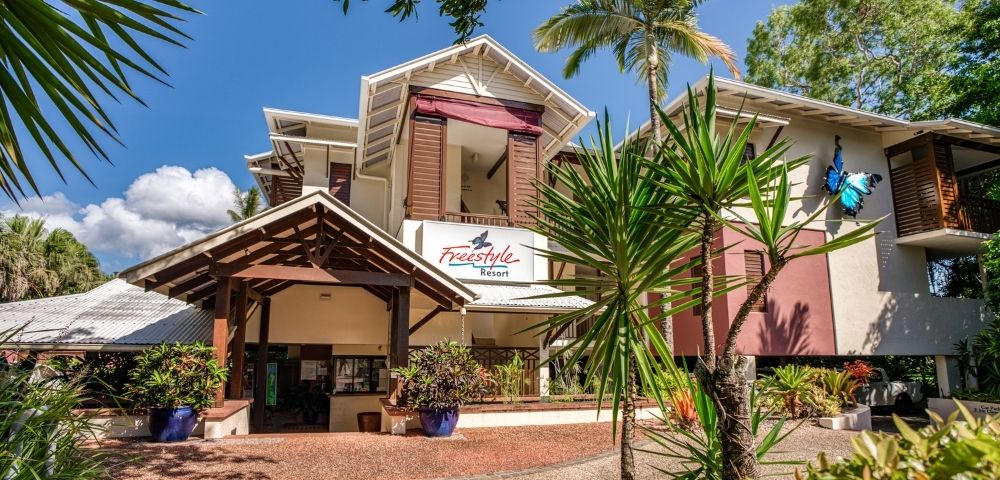 The Freestyle Resort entrance with a covered walkway and tropical plants under a bright blue sky with scattered clouds.