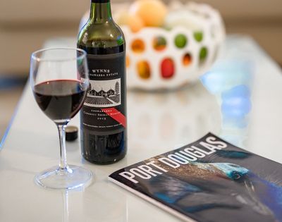 A glass of red wine sits next to a bottle of Wynn's Coonawarra Estate Cabernet Shiraz Merlot 2015 and a magazine titled 