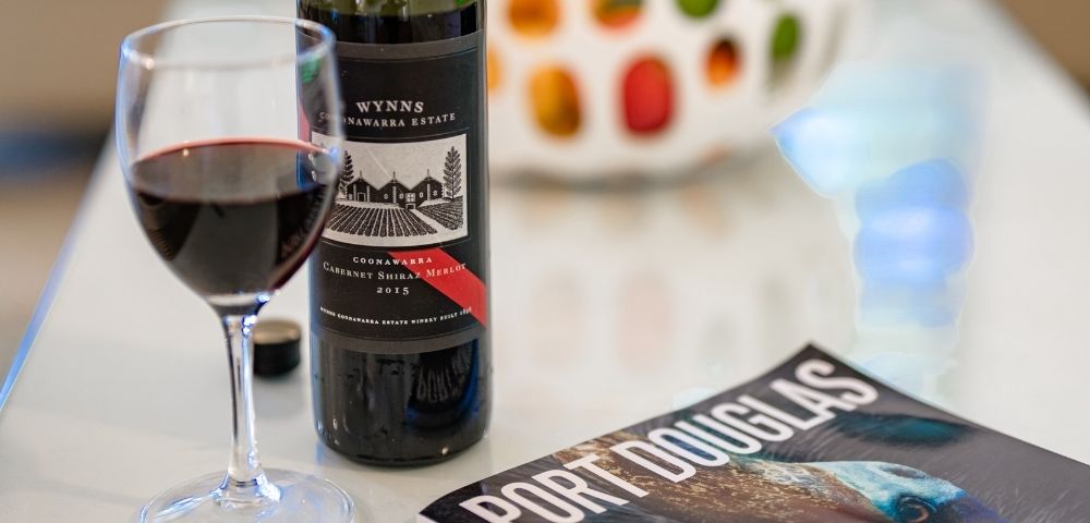 A glass of red wine sits next to a bottle of Wynn's Coonawarra Estate Cabernet Shiraz Merlot 2015 and a magazine titled 