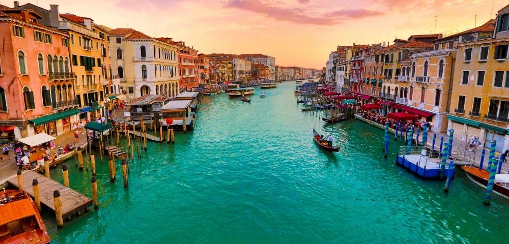 A picturesque canal in Venice at sunset, with vibrant turquoise water, lined with colorful historic buildings and gondolas.