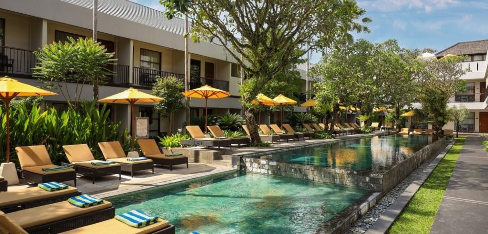 Resort swimming pool with many loungers and yellow umbrellas, surrounded by lush greenery and hotel buildings.