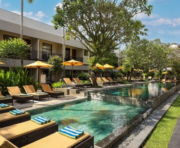 Resort swimming pool with many loungers and yellow umbrellas, surrounded by lush greenery and hotel buildings.