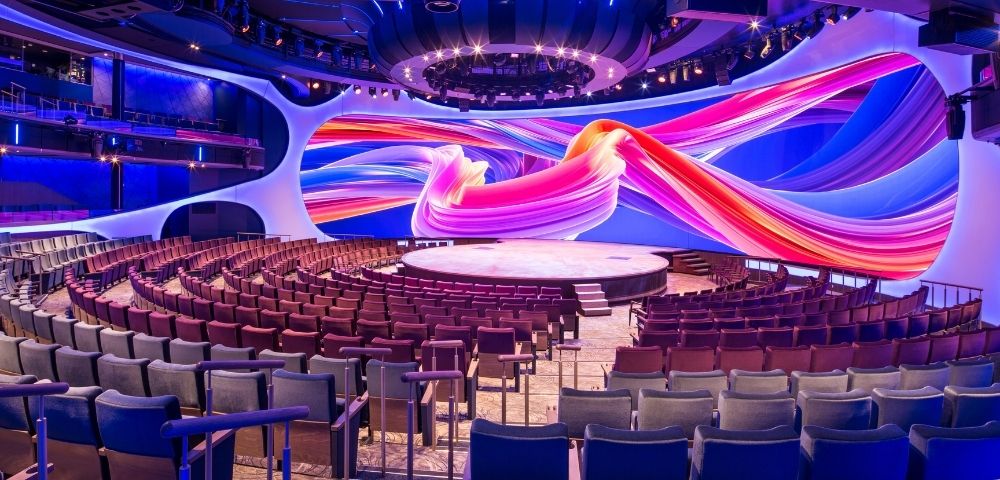 An empty theatre with rows of red and blue seats facing a circular stage under a large, colorful digital screen displaying abstract, swirling shapes in pink, orange, and purple against a blue background.