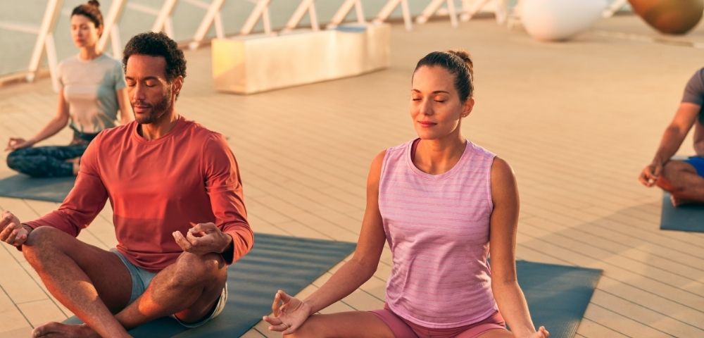 Two people in athletic wear meditate outdoors on yoga mats on a wooden deck, with warm sunlight illuminating the scene.