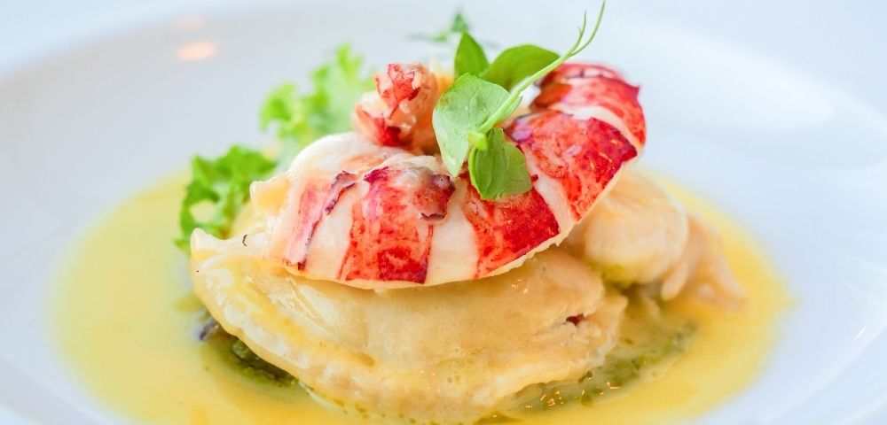 A succulent lobster tail served atop delicate scallops and a bed of green puree in a rich, buttery sauce on a white plate.