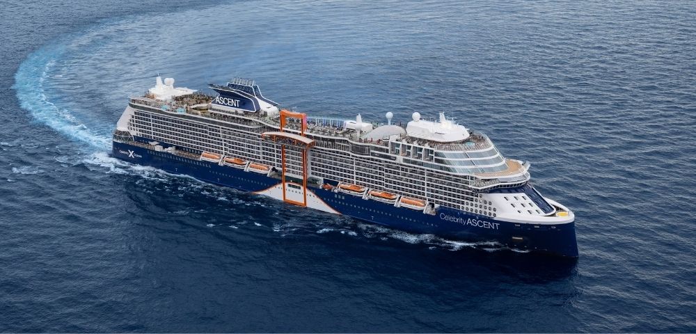 A large blue and white cruise ship, the Celebrity Ascent, sails through dark blue ocean water, leaving a white wake.