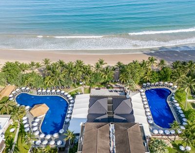 Aerial view of a beachfront resort with two swimming pools, surrounded by palm trees and sun loungers, next to a sandy beach and ocean.