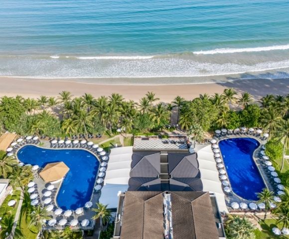 Aerial view of a beachfront resort with two swimming pools, surrounded by palm trees and sun loungers, next to a sandy beach and ocean.