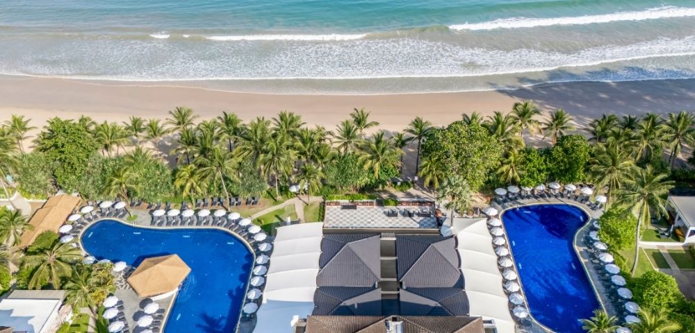 Aerial view of a beachfront resort with two swimming pools, surrounded by palm trees and sun loungers, next to a sandy beach and ocean.