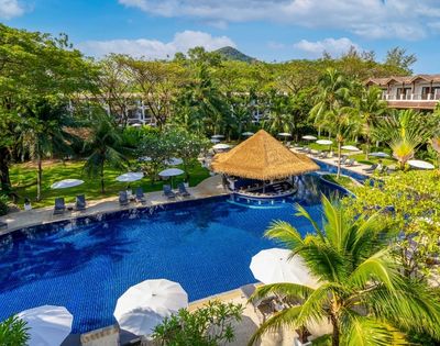 A resort pool with a thatched-roof swim-up bar, surrounded by lounge chairs, umbrellas, palm trees, and lush green hills in the background.
