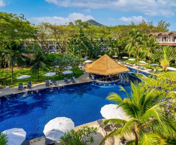 A resort pool with a thatched-roof swim-up bar, surrounded by lounge chairs, umbrellas, palm trees, and lush green hills in the background.