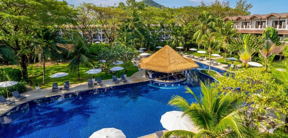 A resort pool with a thatched-roof swim-up bar, surrounded by lounge chairs, umbrellas, palm trees, and lush green hills in the background.